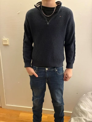 Mörkblå halvzip-tröja Tommy Hilfiger - Mörkblå stickad tröja från Tommy Hilfiger med halvzip och liten logga på bröstet. Tröjan har ribbade muddar vid ärmslut och nederkant samt en hög krage med randig detalj på insidan. Perfekt för en stilren och avslappnad look. Hör av dig för frågor och funderingar!!!