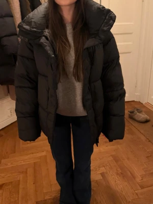 Svart oversized pufferjacka - Säljer en svart oversized pufferjacka med hög krage och breda kanaler. Jackan har en klassisk boxig passform och är perfekt för kalla vinterdagar. Materialet är glansigt och ger en modern look. Dragkedja framtill och stora ärmar för extra komfort.