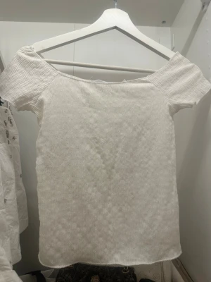 Vit offshoulder topp i ribbad bomull - Trendig vit offshoulder topp med ribbad struktur och korta ärmar. Tillverkad i mjuk bomull som känns skön mot huden. Perfekt att styla med jeans eller kjol för en fräsch look.