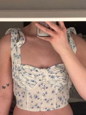 Blommig topp med knytband - Supersöt topp i vitt med blått blommönster och breda axelband som knyts i rosetter. Toppen har en croppad passform, rynkade detaljer framtill och markerad midja. 