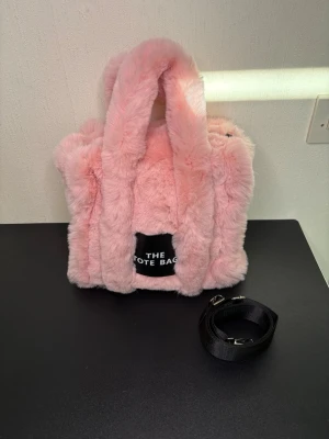 Rosa fluffig handväska med axelrem - Supersöt rosa handväska i mjuk fuskpäls med två handtag och avtagbar svart axelrem. Väskan har en fyrkantig form och en svart patch framtill med texten 'THE TOTE BAG'. Perfekt för dig som vill sticka ut med en mysig accessoar.