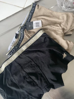 2-pack kalsonger US Grand Polo - Två par kalsonger från US Grand Polo, ett par svarta och ett par beige. Båda har bred resår med logotyp och en liten broderad häst på benet. Snygg och klassisk design, perfekta för vardagsbruk.