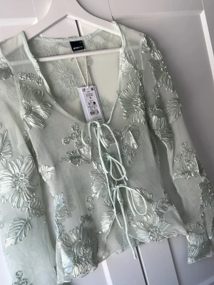 Broderad meshblus från Gina Tricot - Superfin helt ny  ljusgrön blus i mesh från Gina Tricot med broderade blommor och snörning. Blusen har v-ringning och långa ärmar. Blusen är mer åt en ljus mint grön färg än vad som upplevs på bilden 