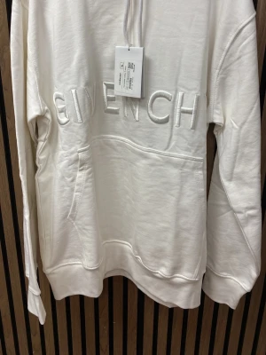 Vit hoodie  - Säljer en stilren vit hoodie med broderad logga framtill. Hoodien har huva med dragsko och en stor magficka. Tillverkad i mjukt material som känns skönt mot huden. Perfekt för dig som vill ha en clean och exklusiv look.