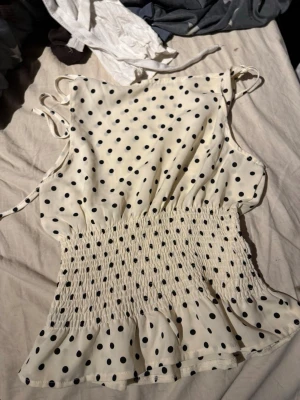 Prickig vit linnetopp med knyt - Söt vit linnetopp med svarta prickar och smockad midja. Toppen har tunna axelband med knyt på axlarna och volangkant nedtill. Perfekt för en trendig och luftig sommarlook.