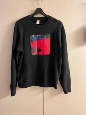 Svart sweatshirt med KAWS-print - Svart sweatshirt från Uniqlo med färgstarkt KAWS-motiv på bröstet i rött och blått. Baksidan har broderad text och två vita kryss samt ett tryckt fotomotiv på nedre delen. Rund hals och avslappnad passform, perfekt för streetwear.
