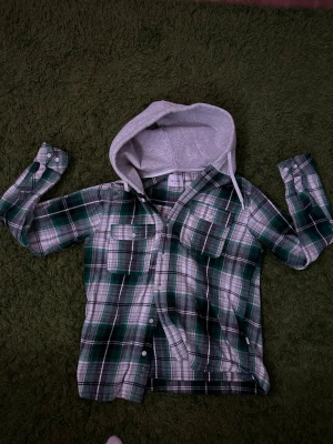 Grönrutig overshirt med huva från Vaillent - Säljer en grön, vit och grå rutig overshirt från Vaillent. Skjortan har en grå huva med dragsko, två bröstfickor med knappar och långa ärmar. Perfekt lager på lager-plagg med avslappnad vibe och skön passform. Helt oanvänd, går för 600kr ny