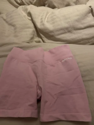 Rosa seamless shorts från DFYNE - Säljer ett par ljusrosa seamless shorts från DFYNE med hög midja och ribbad linning. Shortsen har en scrunch-detalj bak och är tillverkade i ett stretchigt, mjukt material som passar perfekt till träning eller chill. Loggan sitter diskret framtill i vitt.