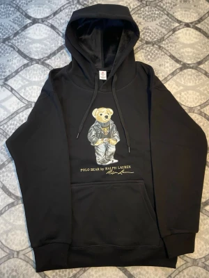 Svart hoodie Polo Bear Ralph Lauren - Svart hoodie från Ralph Lauren med den ikoniska Polo Bear-trycket på bröstet. Klassisk känguruficka framtill, dragsko i huvan och ribbade muddar. Tillverkad i mjukt bomullsmaterial för skön känsla. Perfekt för en avslappnad och trendig stil.