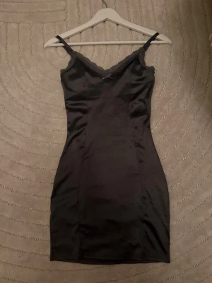 Mörk grå satin klänning med spets - Snygg mörk grå kortklänning i satin med smala axelband och spetsdetaljer vid urringningen. Figurnära passform och enkel, stilren design som ger en elegant look. Perfekt för dig som vill ha en klassisk och trendig klänning.