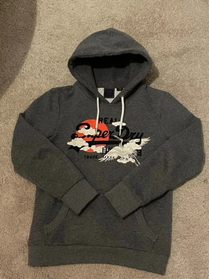 Mörkgrå Superdry hoodie med tryck - Säljer en mörkgrå hoodie från Superdry med stort grafiskt tryck i rött, vitt och svart framtill. Hoodien har huva med vita dragsnören och en klassisk känguruficka. Perfekt för dig som gillar streetwear och vill ha en snygg och bekväm stil.