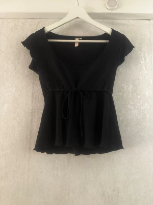 Svart topp med knyt och volang - Säljer en svart topp med korta ärmar och rundad urringning. Toppen har en söt volangkant nedtill och knytband i midjan för en feminin siluett. Materialet är ribbat och stretchigt, vilket gör den bekväm och smickrande.