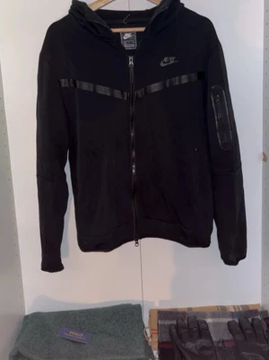 🔥 ÄKTA Nike Tech Hoodie – Svart (Storlek L) - Säljer en original Nike Tech hoodie i klassisk svart färg, storlek L. Hoodien är lite använd men i bra skick, inga större slitningar. Perfekt för vardag, skola eller streetwear-look.  ✔️ Äkta Nike ✔️ Storlek L ✔️ Svart ✔️ Lite använd – väl omhändertagen  Hör av dig vid intresse eller om du vill ha fler bilder 😊