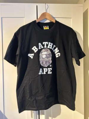 Svart A Bathing Ape t-shirt - Säljer en svart t-shirt från A Bathing Ape med det ikoniska BAPE-huvudet i camomönster och stor vit text på bröstet. Klassisk rund hals och korta ärmar. Perfekt för dig som gillar streetwear och vill sticka ut med en hypead tee. Helt ny priset kan diskuteras 