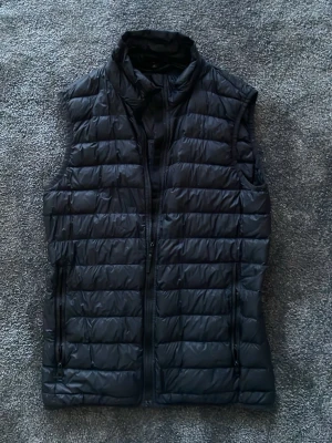 Svart dunväst J Lindeberg - Snygg svart dunväst med horisontella sömmar och hög krage. Västen har två fickor med dragkedja och stängs med en smidig dragkedja framtill. Perfekt att slänga över en hoodie eller tröja för extra värme. Tillverkad i lätt och mjukt material.