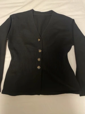 Svart ribbad kofta med knappar - Säljer en svart kofta med ribbade partier i midjan och fyra dekorativa knappar framtill. Koftan har V-ringning och långa ärmar, perfekt för lager på lager. Materialet är mjukt och stretchigt för en bekväm passform.
