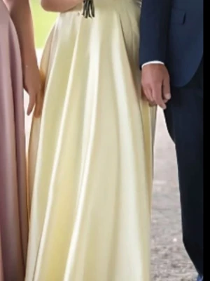 Ljusgul långklänning i satin - Elegant ljusgul långklänning med omlottdetalj upptill och markerad midja. Klänningen har en vid, svepande kjol och är ärmlös, vilket ger en stilren och klassisk look. Perfekt för bal eller student. Ligger på flera sidor.                               Originalpåse finns!!! 