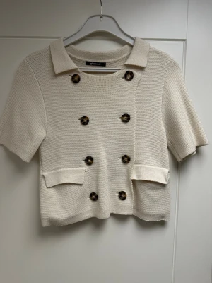 Krämvit kofta med kort ärm från Gina Tricot S - Stilren krämvit kofta från Gina Tricot med korta ärmar, dubbelknäppning med stora bruna knappar och två dekorativa fickor framtill. Koftan har en klassisk krage och är stickad i ett mjukt material. Perfekt för en chic och trendig look.
