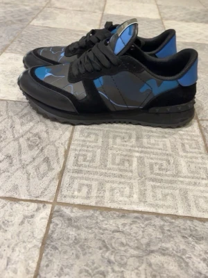Valentino rockrunners - Snygga sneakers med svart bas och blått camomönster. Skorna har svarta snören, svart sula med nitar och detaljer i mocka och läder. Insidan är vadderad för extra komfort och hälen har en blå kontrastpanel. Använda bara en gång köpta helt nya från hemsidan