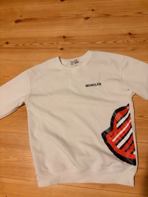 Vit sweatshirt från Moncler - Säljer en vit sweatshirt från Moncler med rund halsringning och lång ärm. Tröjan har en stor Moncler-logga i rött, svart och vitt på sidan samt diskret Moncler-tryck på bröstet. Perfekt för dig som gillar stilrena plagg med en twist.