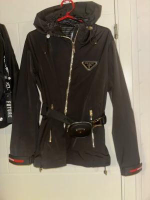 Svart jacka från Prada med bälte storlekS/M - Snygg svart  från Prada med huva, silvriga dragkedjor och justerbart bälte i midjan. Jackan har en liten väska på bältet med Prada-logga och dragsko i nederkant. Aldrig använd, ny.
