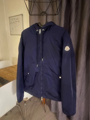 Mörkblå vindjacka från Moncler - Snygg mörkblå vindjacka från Moncler med huva och dragkedja framtill. Jackan har två fickor med tryckknappar och Moncler-logga på ärmen. Perfekt för blåsiga dagar och enkel att matcha med olika outfits.
