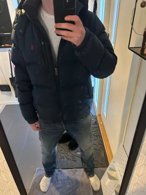  pufferjacka från Ralph Lauren - Säljer en marinblå pufferjacka från Ralph Lauren med röd logga på bröstet. Jackan har dragkedja framtill, jackan har några små defekter som man ser på bilden. Priset är ej hugget i sten