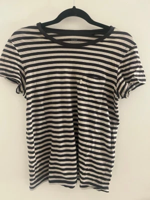 Randig svartvit t-shirt med ficka - Säljer en svartvit randig t-shirt med rund hals och en bröstficka. T-shirten har korta ärmar och är tillverkad i mjuk bomull, perfekt för en avslappnad stil. Passar dig som gillar klassiska mönster och enkel design.