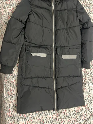 Svart lång dunjacka med reflexdetaljer - Snygg svart lång dunjacka med dragkedja framtill och två stora fickor med reflexdetaljer. Jackan har ribbade muddar vid ärmsluten och är perfekt för kalla vinterdagar. Materialet är glansigt och quiltat för extra värme.