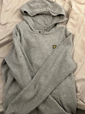Grå hoodie från Lyle & Scott - Säljer en stilren grå hoodie från Lyle & Scott med deras klassiska gula fågel-logga broderad på bröstet. Hoodien har huva med dragsko och en stor magficka. Tillverkad i mjuk bomullsblandning och perfekt för en avslappnad look. Står i storleken att den passar 14-15 år men jag skulle säga att det är S.
