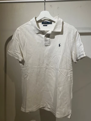 Vit pikétröja från Polo Ralph Lauren - Klassisk vit pikétröja från Polo Ralph Lauren med blå broderad logga på bröstet. Slim fit-modell med korta ärmar och krage. Tillverkad i bomull för en skön och luftig känsla. Perfekt till jeans eller shorts för en clean look.(XL i barn så passar S)