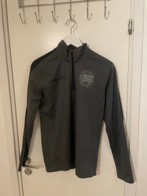 Hammarby träningströja  - Mörkgrå långärmad träningsjacka från Craft med halv dragkedja framtill och tryckt bajen-logga på bröstet. Jackan har hög krage och svarta detaljer på axlarna. Perfekt för löpning eller träning utomhus.