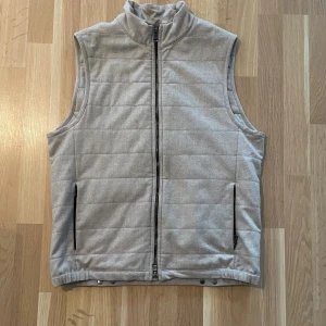 Grå väst från Oscar Jacobson - Snygg grå väst från Oscar Jacobson. Västen har två fickor med dragkedja och stilrena tryckknappar nedtill. Perfekt att slänga på över en hoodie eller tröja när det är lite kyligt ute. Går att sänka priset vid snabb affär