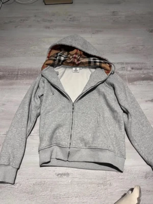 Grå zip-hoodie från Burberry - Snygg grå hoodie från Burberry med dragkedja och klassiskt rutigt foder i huvan. Mjuk insida och ribbade muddar vid ärmslut och nederkant. Perfekt för en avslappnad och stilren look. Helt ny och oanvänd!! 