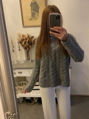 Grå kabelstickad tröja - Mysig grå kabelstickad tröja med rund hals och långa ärmar. Tröjan har ett klassiskt flätmönster och är lite oversized i modellen, perfekt för lager-på-lager. Materialet ser ut att vara mjukt och värmande, passar bra till både jeans och mjukisbyxor.