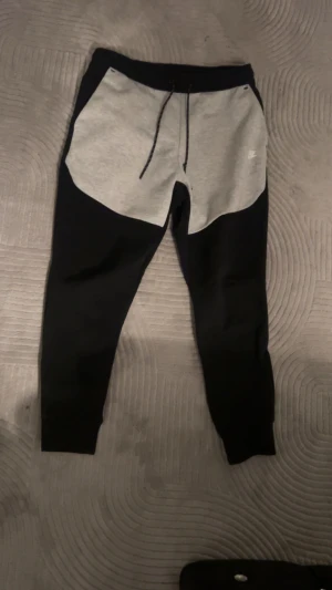 Nike tech byxor - Säljer ett par snygga mjukisbyxor från Nike i svart och grått. Byxorna har dragsko i midjan och klassisk Nike-logga på ena sidan. De är tillverkade i mjukt material och har en modern, avsmalnande passform som är perfekt för chill eller träning.