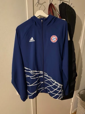 Blå FC Bayern München hoodie Adidas - Snygg blå hoodie från Adidas med FC Bayern München-logga på bröstet och vit text på ryggen. Jackan har hel dragkedja, huva och vita grafiska linjer nedtill. Perfekt för dig som älskar fotboll och vill ha en sportig look.