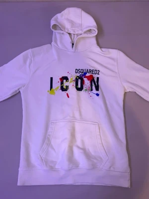 Vit hoodie DSQUARED2 ICON - Säljer en vit hoodie från DSQUARED2 med färgglad ICON-print och färgstänk på bröstet. Klassisk känguruficka framtill och huva. Perfekt för dig som vill sticka ut med en streetstyle-look.