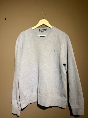 Ljusblå tröja från Polo Ralph Lauren - Klassisk ljusblå tröja från Polo Ralph Lauren i 100% bomull. Tröjan har V-ringning, långa ärmar och den ikoniska broderade loggan på bröstet. Perfekt för en clean och stilren look.