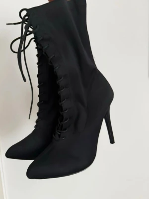 Svarta stövletter med snörning - Snygga svarta stövletter med hög smal klack och spetsig tå. Skorna har snörning framtill som ger en edgy look och är tillverkade i ett slätt syntetmaterial. Perfekta för dig som vill sticka ut med en stilren och trendig sko. Sömnen där bak ör lite av men inget som påverkar