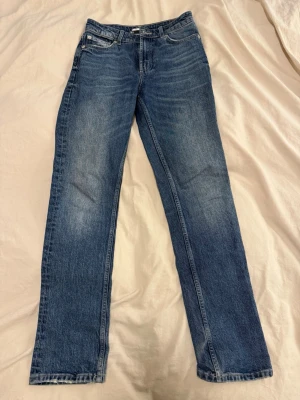 Blå raka jeans med klassisk tvätt - Säljer ett par blå jeans med rak passform och klassisk tvätt. Jeansen har fem fickor, bälteshällor och subtila slitningar för en avslappnad look. Perfekta till vardags och passar till det mesta. Materialet är denim i bomull.
