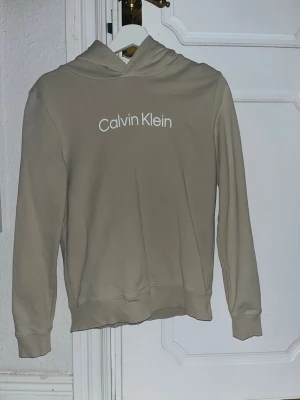 Beige hoodie från Calvin Klein - Snygg beige hoodie från Calvin Klein med vit logga tryckt på bröstet. Klassisk modell med huva och ribbade muddar vid ärmar och nederkant. Tillverkad i mjukt material som känns skönt mot huden. Perfekt för en avslappnad och stilren look.