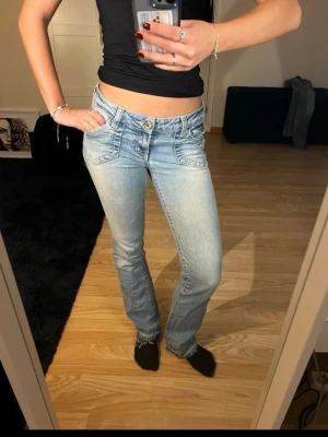 Ljusblå bootcut jeans med låg midja - Säljer ett par ljusblå bootcut jeans med låg midja och snygga slitningar. Jeansen har framfickor med detaljerade sömmar och en klassisk knappgylf. Perfekta för en avslappnad och trendig look. De har lite hål där och där, har sprättat upp längst ner för att de ska bli längre och även ett snöre på insidan!🤗🤗