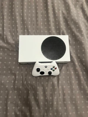 Xbox Series S med handkontroll - Säljer en Xbox Series S i vit färg med tillhörande vit trådlös handkontroll. Konsolen har en kompakt, rektangulär design med en stor svart cirkulär ventil på ovansidan. Perfekt för gaming och digitala spel. Materialet är plast. Hämtas i Södertälje 