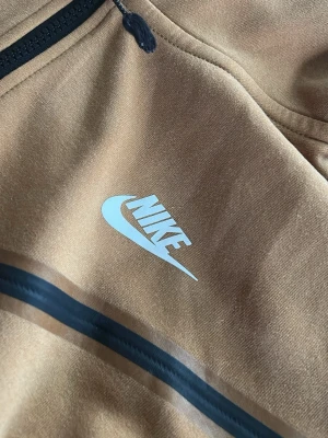 Brun Nike hoodie med dragkedja - Snygg brun hoodie från Nike med hel dragkedja framtill och svart detalj längs bröstet. Hoodien har en stor huva och Nike-logga i vitt på bröstet. Tillverkad i mjukt material som känns skönt mot huden. Perfekt för en avslappnad och sportig stil.