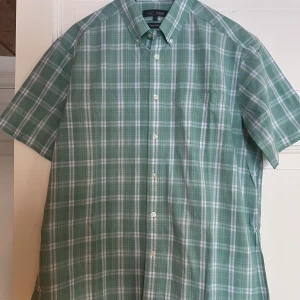 Grön rutig kortärmad skjorta M&S - Kortärmad skjorta från Mark & Spencer, M&S Man i grön och vit rutig bomull. Klassisk krage med knappar och knappstängning framtill. Perfekt för en avslappnad och stilren look.