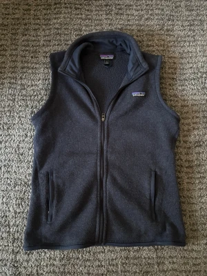 Mörkblå fleeceväst från Patagonia - Mörkblå fleeceväst från Patagonia med hel dragkedja framtill och två fickor med dragkedja. Västen har hög krage och är tillverkad i mjukt fleecematerial. Perfekt för lager-på-lager och outdoor-stil. 