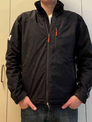 Svart vindjacka från Helly Hansen - Snygg svart vindjacka från Helly Hansen med hög krage och vita HH-loggor på krage och ärm. Jackan har tre fickor med dragkedjor och röda detaljer. Perfekt för blåsiga dagar och enkel att matcha med olika outfits. Längd 66cm, P2P: 51cm. Behöver en tvätt