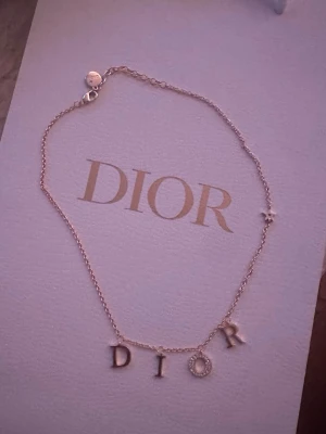 Dior halsband guld  - Snyggt halsband från Dior i guldtonad metall med hängande bokstäver som stavar DIOR. O:et är dekorerat med små gnistrande stenar. Kedjan är tunn och har en liten stjärndetalj på sidan. Perfekt för dig som gillar exklusiva accessoarer.