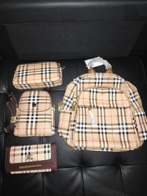 Väskor set  - Säljer ett set med beige och bruna handväskor i klassiskt rutigt mönster. Setet innehåller en handväska, en plånbok, en axelväska och en ryggsäck. Alla delar har guldfärgade detaljer och är tillverkade i syntetmaterial. Perfekt för dig som gillar stilrena accessoarer med en twist. Handväskor: 250 kr st. Ryggsäck 500 kr. Paketpris för allt: 1000 kr. 